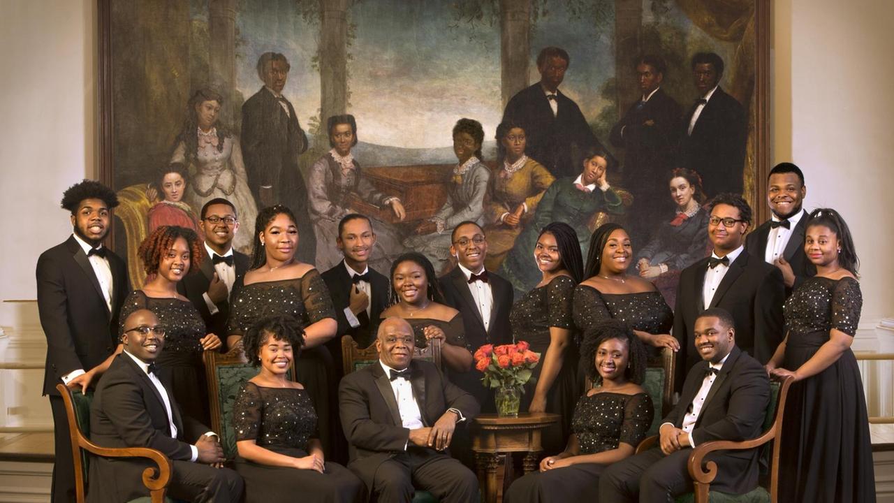Knapp 20 Menschen in schwarzer Konzertkleidung stehen und sitzen vor einem großformatigem Gemälde, das Vorgänger des Chores zeigt, die in historischer Kleidung in einer arkadischen Landschaft beieinander stehen und sitzen.