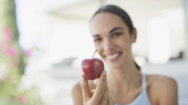 Eine junge Frau hält lachend einen kleinen roten Apfel in den Vordergrund.