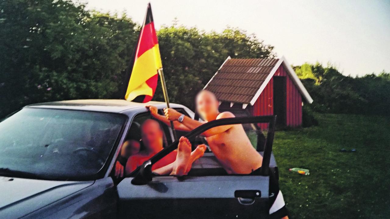 Zwei Männer mit kahlrasierten Schädeln und freien Oberkörpern; der eine sitzt mit ausgestreckten Beinen auf dem Fahrersitz eines Autor, der andere lehnt daran und hält eine Deutschlandfahne; im Hintergrund sieht man noch eine dritte Person. Die Gesichter der drei Männer sind anonymisiert Zwei Männer mit kahlrasierten Schädeln und freien Oberkörpern; der eine sitzt mit ausgestreckten Beinen auf dem Fahrersitz eines Autor, der andere lehnt daran und hält eine Deutschlandfahne; im Hintergrund sieht man noch eine dritte Person. Die Gesichter der drei Männer sind anonymisiert