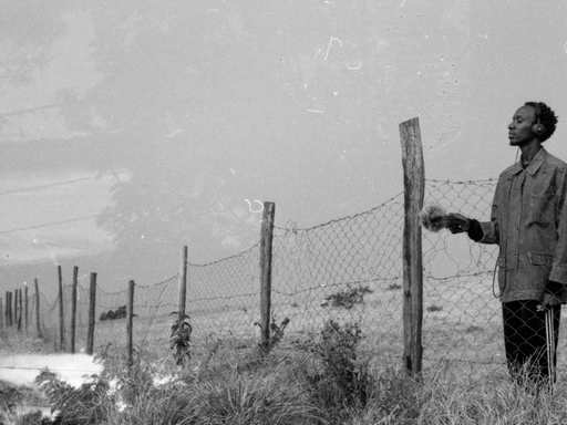 Schwarz-Weiss Fotografie von Joseph Kamaru, der mit Kopfhörern an einem Maschendrahtzaun steht.