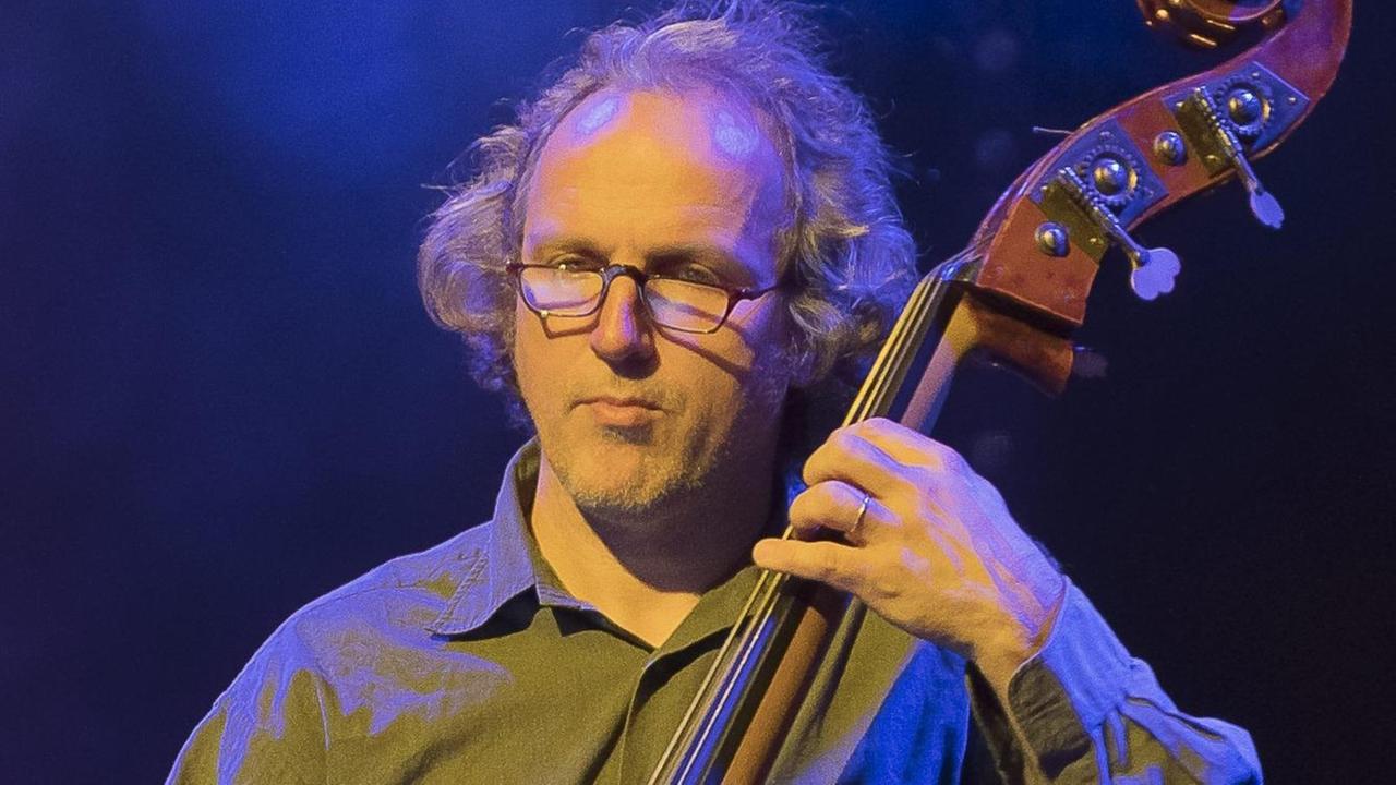 Ein Mann mit Brille greift die Saiten eines Kontrabasses. Er steht blau angeleuchtet vor einem dunklen Bühnen-Hintergrund. Ein Mann mit Brille greift die Saiten eines Kontrabasses. Er steht blau angeleuchtet vor einem dunklen Bühnen-Hintergrund.
