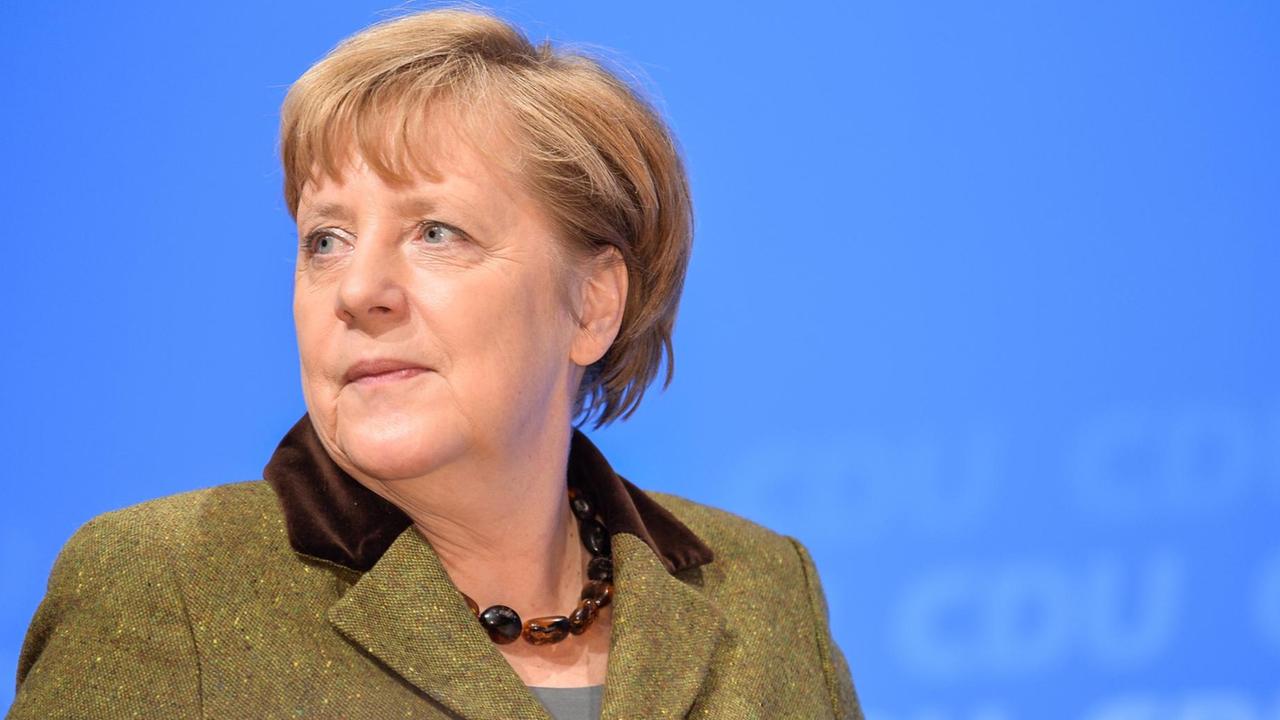 Merkel blickt vor einer blauen Wand mit CDU-Aufdrucken milde lächelnd zur Seite. Sie trägt ein grünes jackett und eine Bernsteinkette.