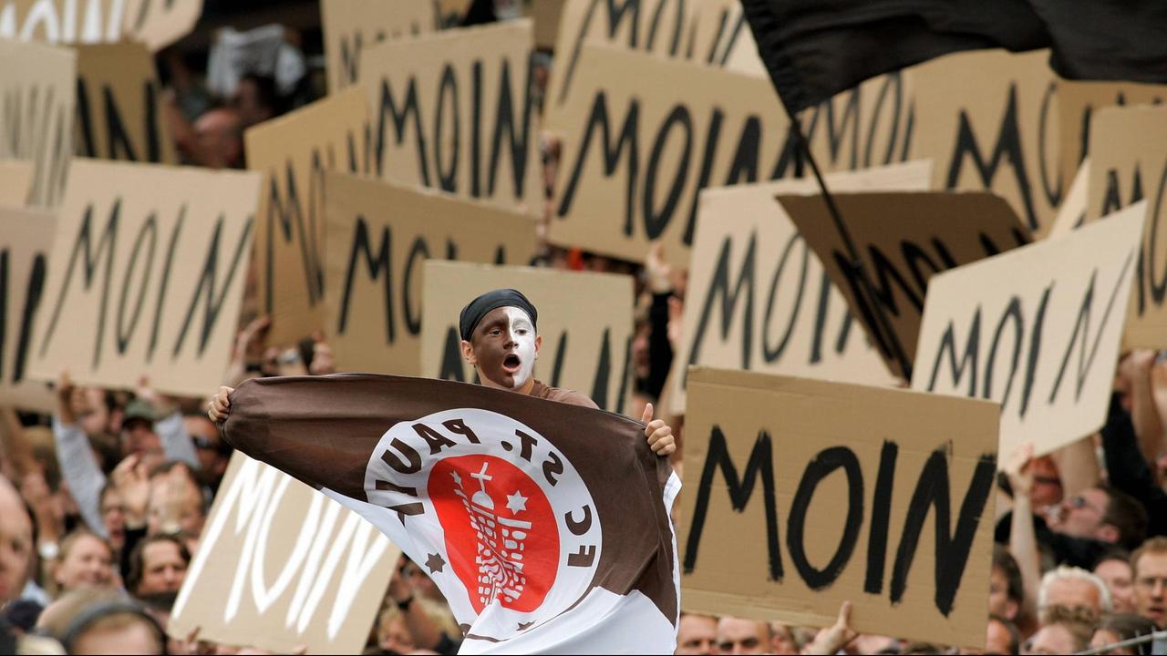 St. Pauli Fan mit Gesichtsbemalung und Vereinsfahne umringt von Fanplakaten auf denen "Moin" steht. St. Pauli Fan mit Gesichtsbemalung und Vereinsfahne umringt von Fanplakaten auf denen "Moin" steht.