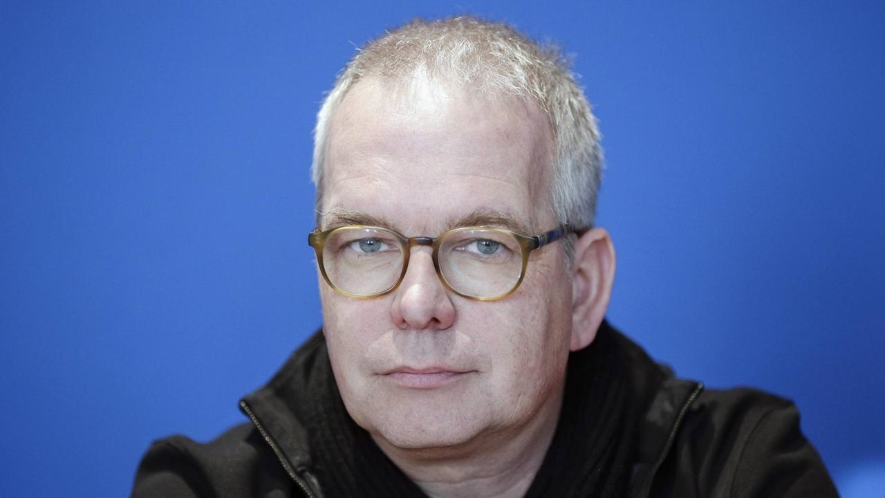 Ein Mann mit etwas schütterem Haar und Brille blickt vor einem blauen Hintergrund in die Kamera. Ein Mann mit etwas schütterem Haar und Brille blickt vor einem blauen Hintergrund in die Kamera.
