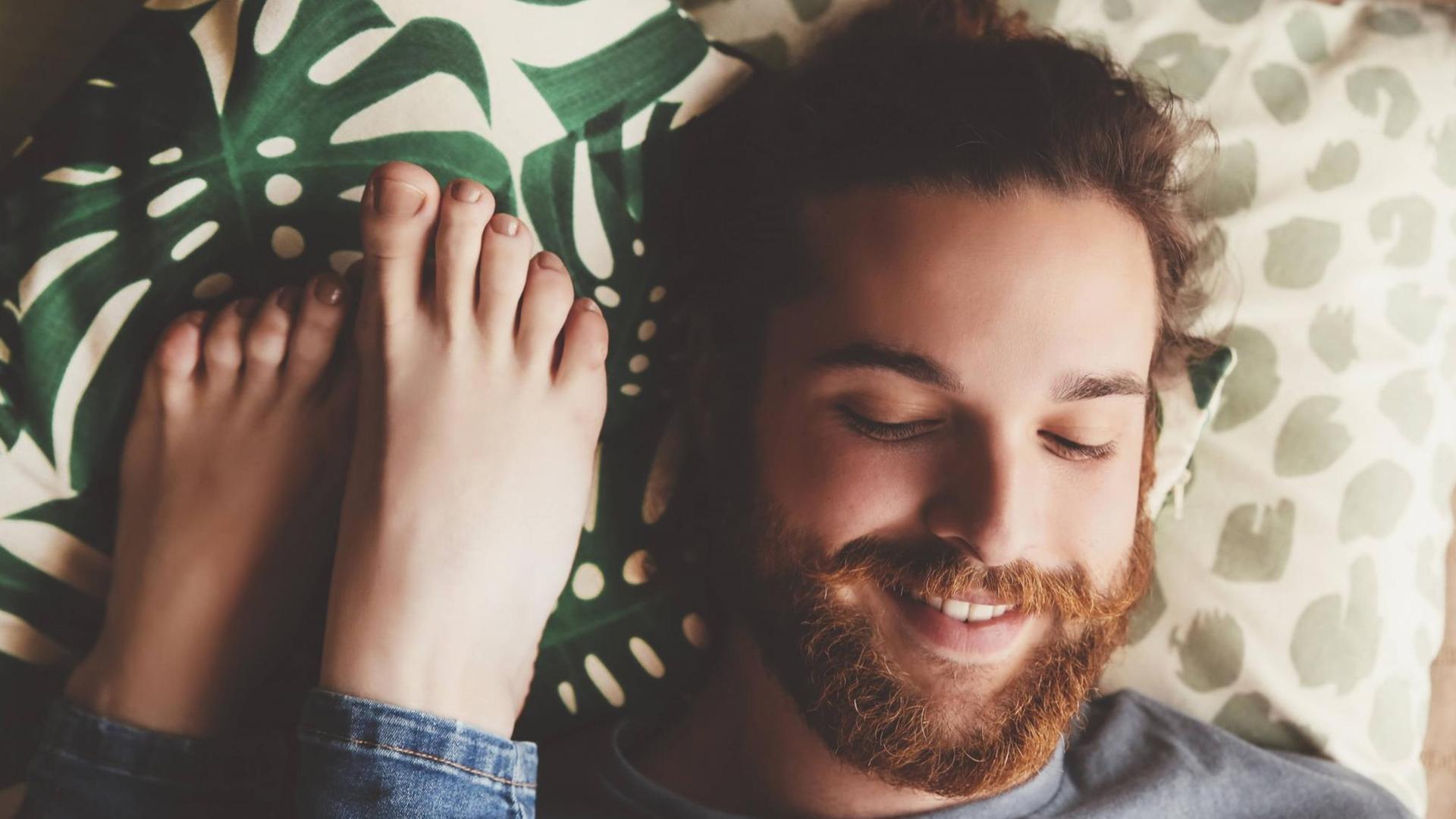 Ein lächelnder Mann liegt auf dem Bett, neben ihm Füße einer Frau. Ein lächelnder Mann liegt auf dem Bett, neben ihm Füße einer Frau.