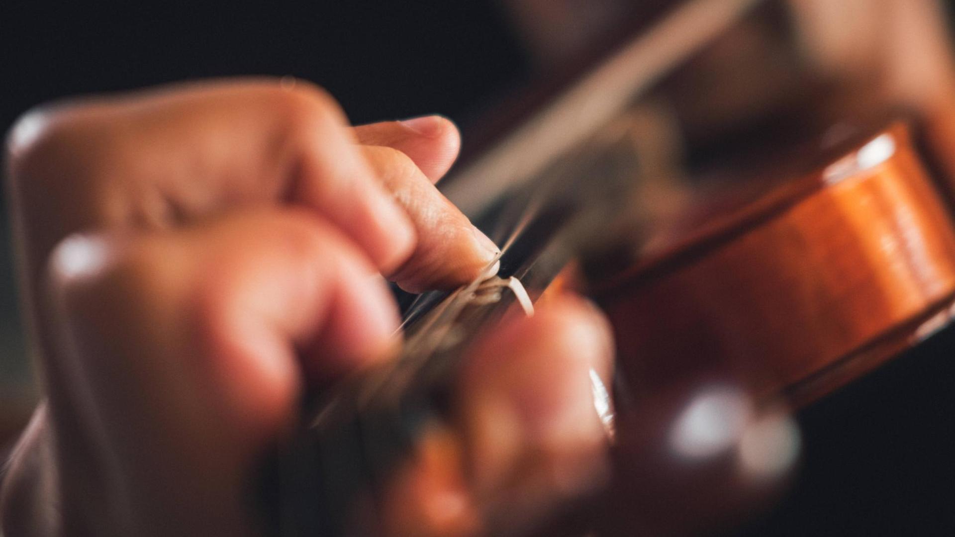 Im Vordergrund ist eine Hand mit aufgestellten Fingern auf einem Geigengriffbrett zu sehen. Im Vordergrund ist eine Hand mit aufgestellten Fingern auf einem Geigengriffbrett zu sehen.