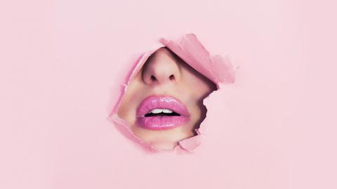 Durch ein Loch in einer rosa Pappe ist ein pink geschminkter Mund und die Nase einer Frau zu sehen.