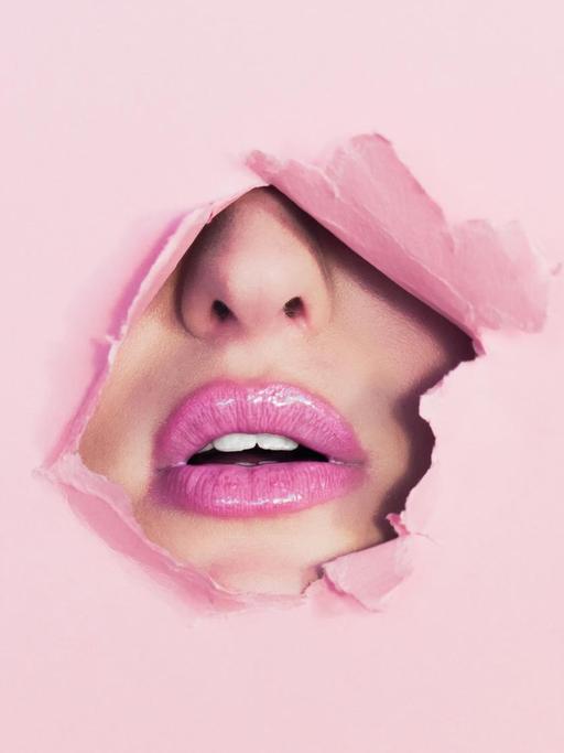 Durch ein Loch in einer rosa Pappe ist ein pink geschminkter Mund und die Nase einer Frau zu sehen.