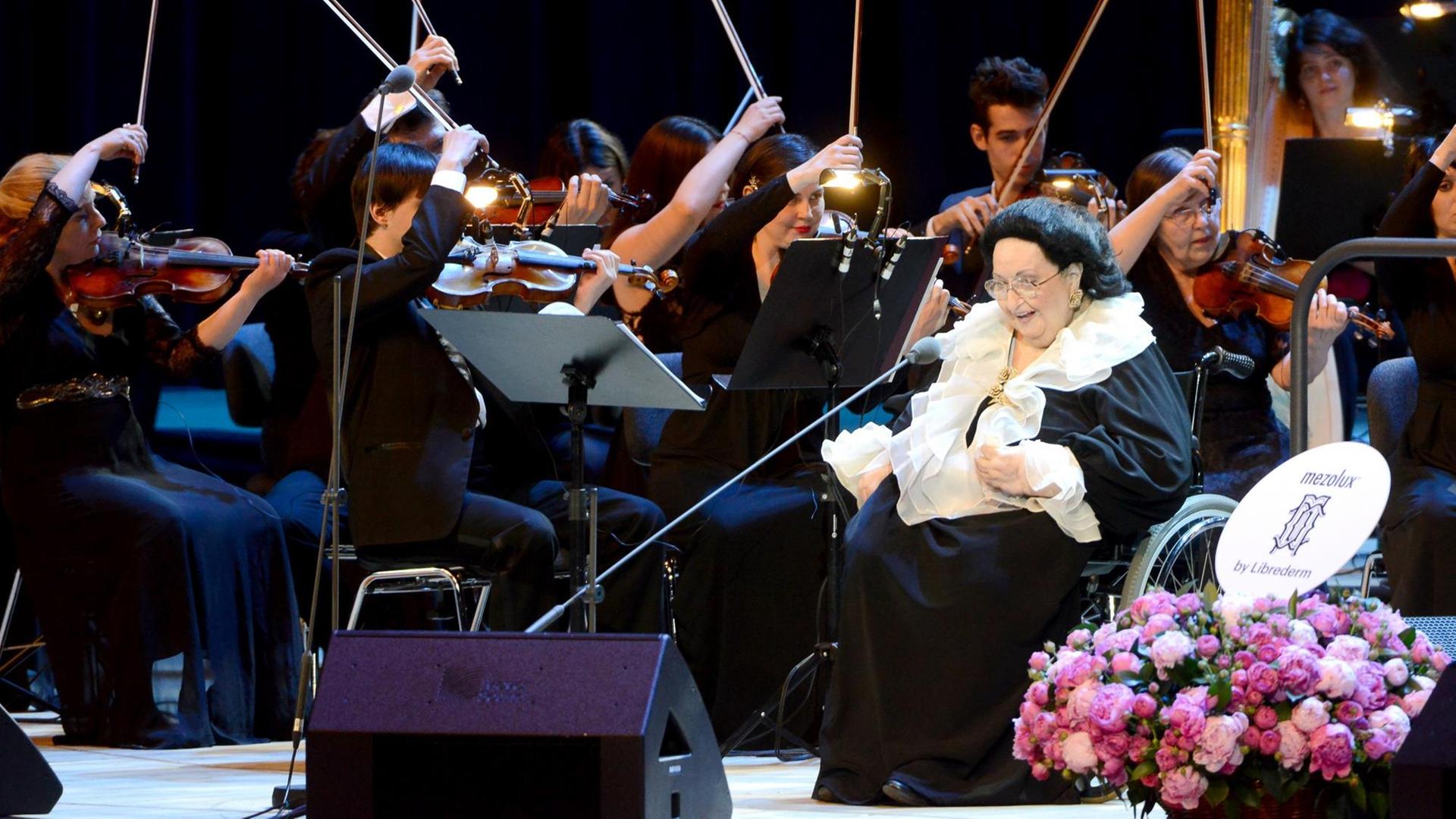 Montserrat Caballé sitzt in einem Rollstuhl auf der Bühne, hinter ihr Teile des Orchesters.