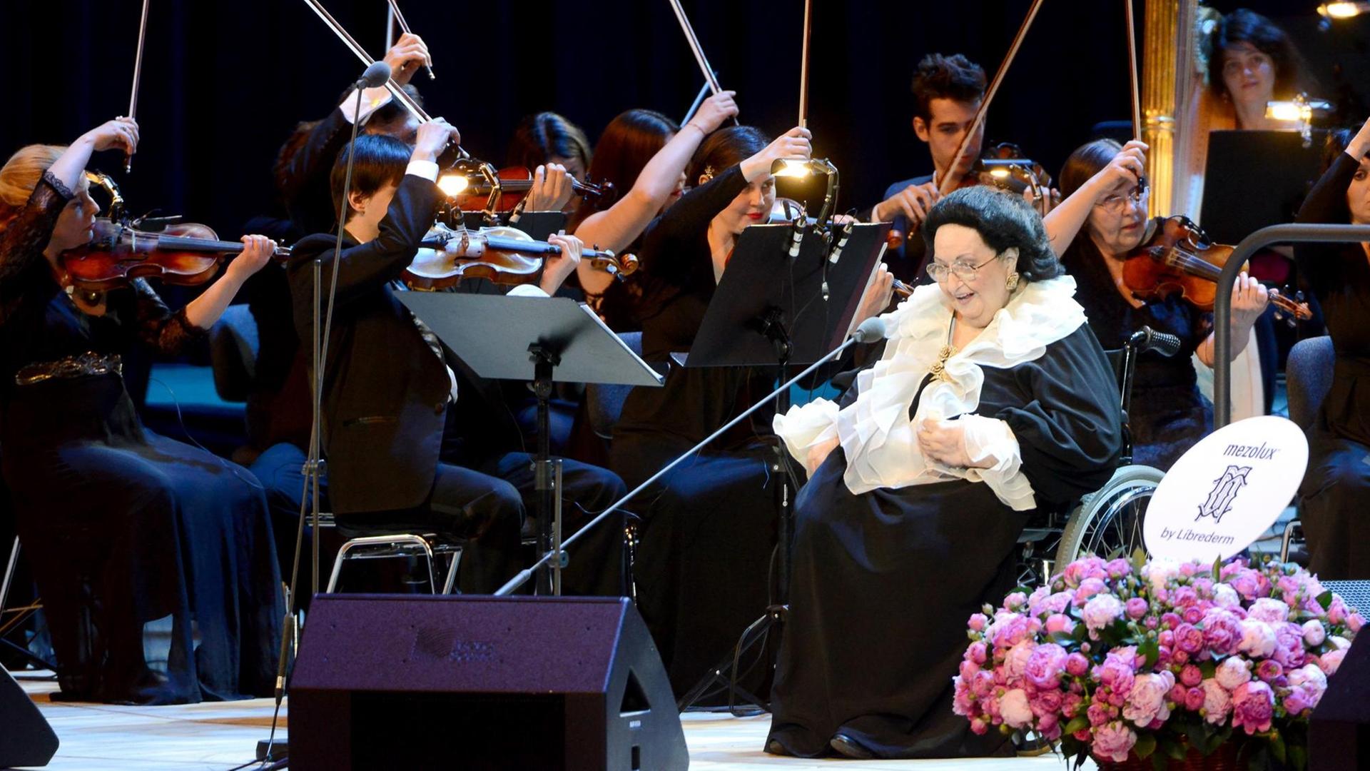 Montserrat Caballé sitzt in einem Rollstuhl auf der Bühne, hinter ihr Teile des Orchesters. Montserrat Caballé sitzt in einem Rollstuhl auf der Bühne, hinter ihr Teile des Orchesters.