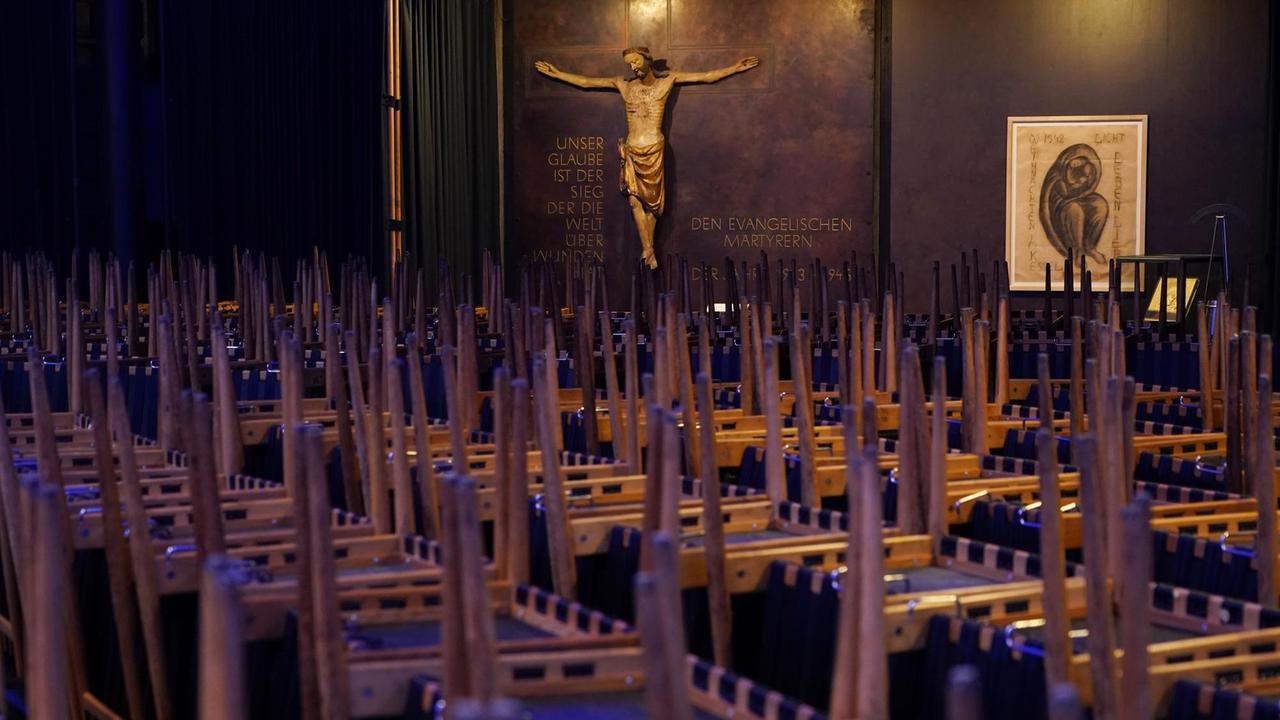 Die Stühle in einer Kirche sind allesamt hochgestellt, im Hintergrund ist eine Jesus-Figur an der Wand zu sehen. Die Stühle in einer Kirche sind allesamt hochgestellt, im Hintergrund ist eine Jesus-Figur an der Wand zu sehen.