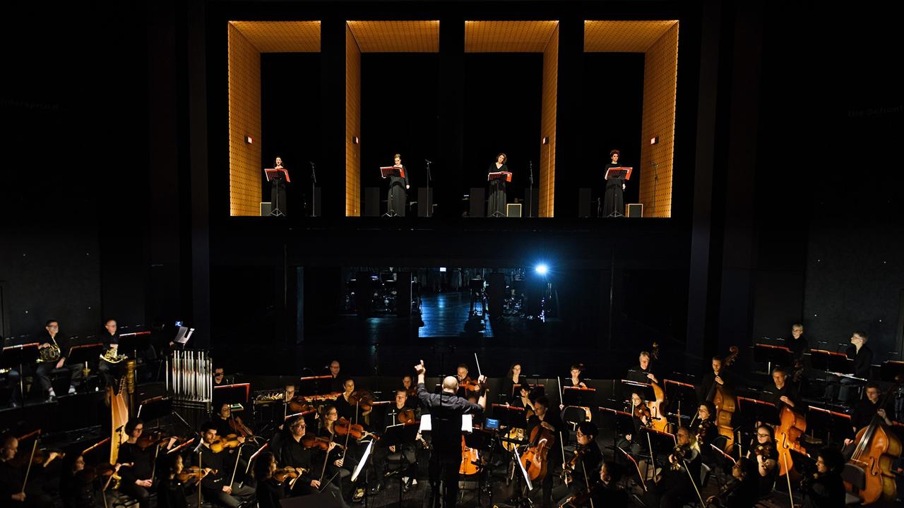 Das Orchester im erhobenen Orchestergraben und Solisten auf der Bühne sind gleichermaßen sichtbar. Das Orchester im erhobenen Orchestergraben und Solisten auf der Bühne sind gleichermaßen sichtbar.