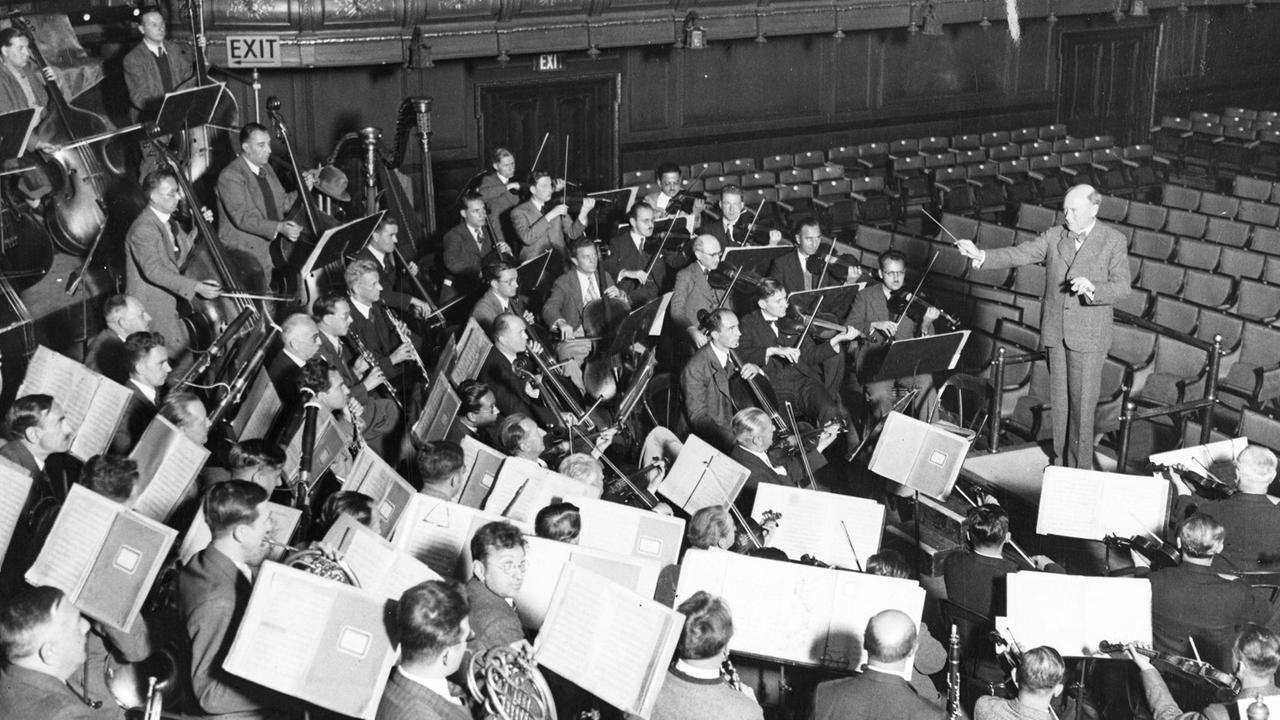 Auf einer historischen Fotografie ist ein Dirigent zu sehen, der ein sehr dicht besetztes Orchester mit großen Notenpulten leitet.