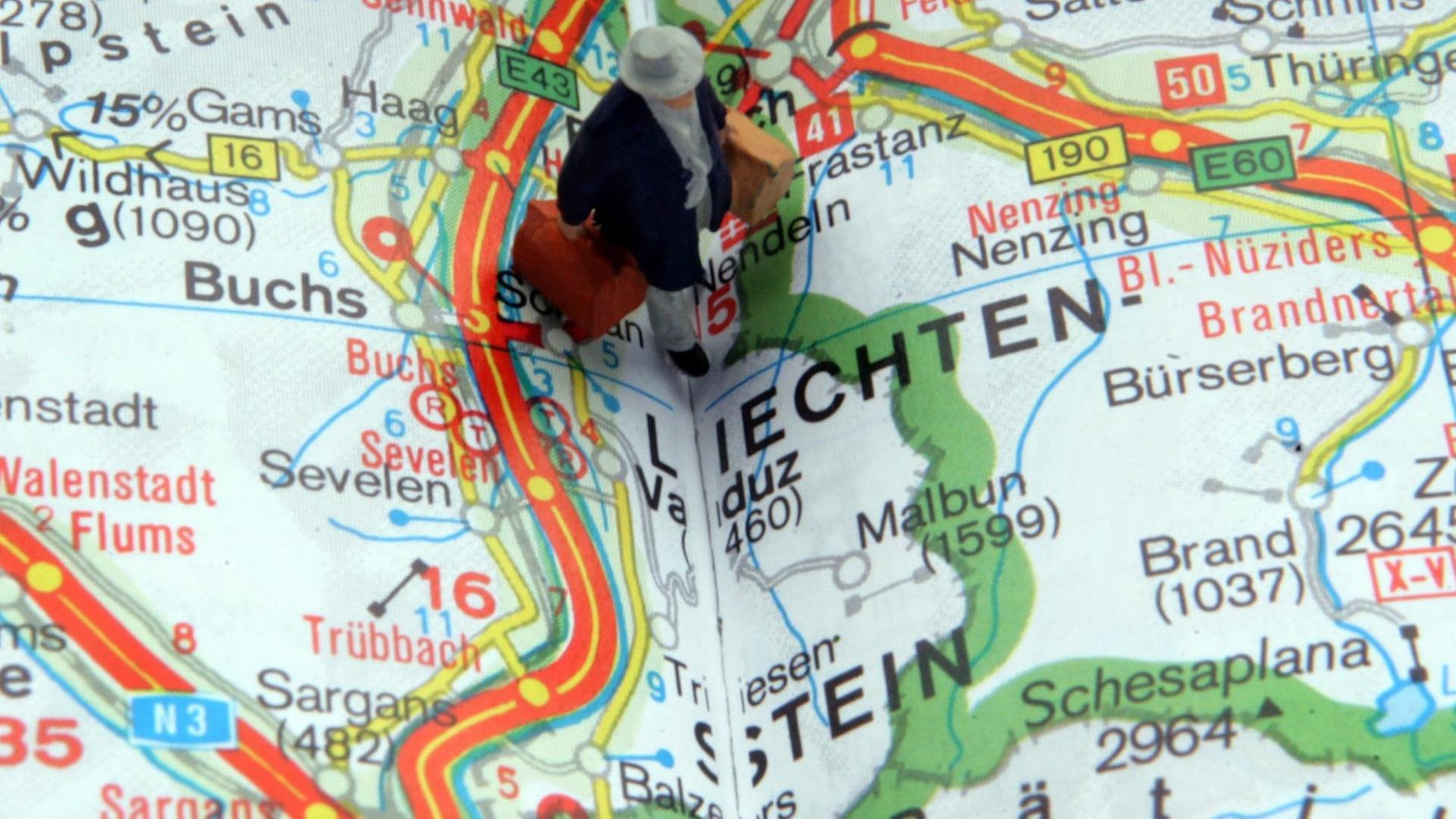 Eine Miniaturfigur mit zwei Koffer steht auf einer Landkarte an der Grenze zu Liechtenstein ( Eine Miniaturfigur mit zwei Koffer steht auf einer Landkarte an der Grenze zu Liechtenstein (