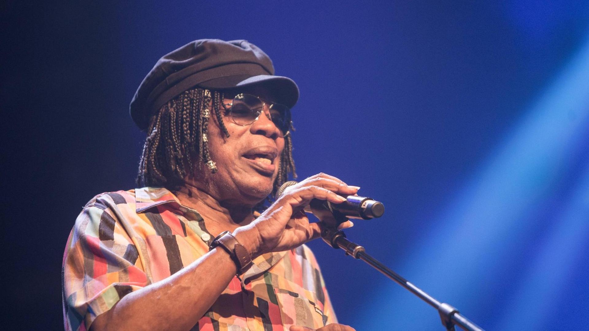 Milton Nascimento trägt eine Brille und eine Mütze und singt auf einer Bühne in ein Mikrofon. Milton Nascimento trägt eine Brille und eine Mütze und singt auf einer Bühne in ein Mikrofon.