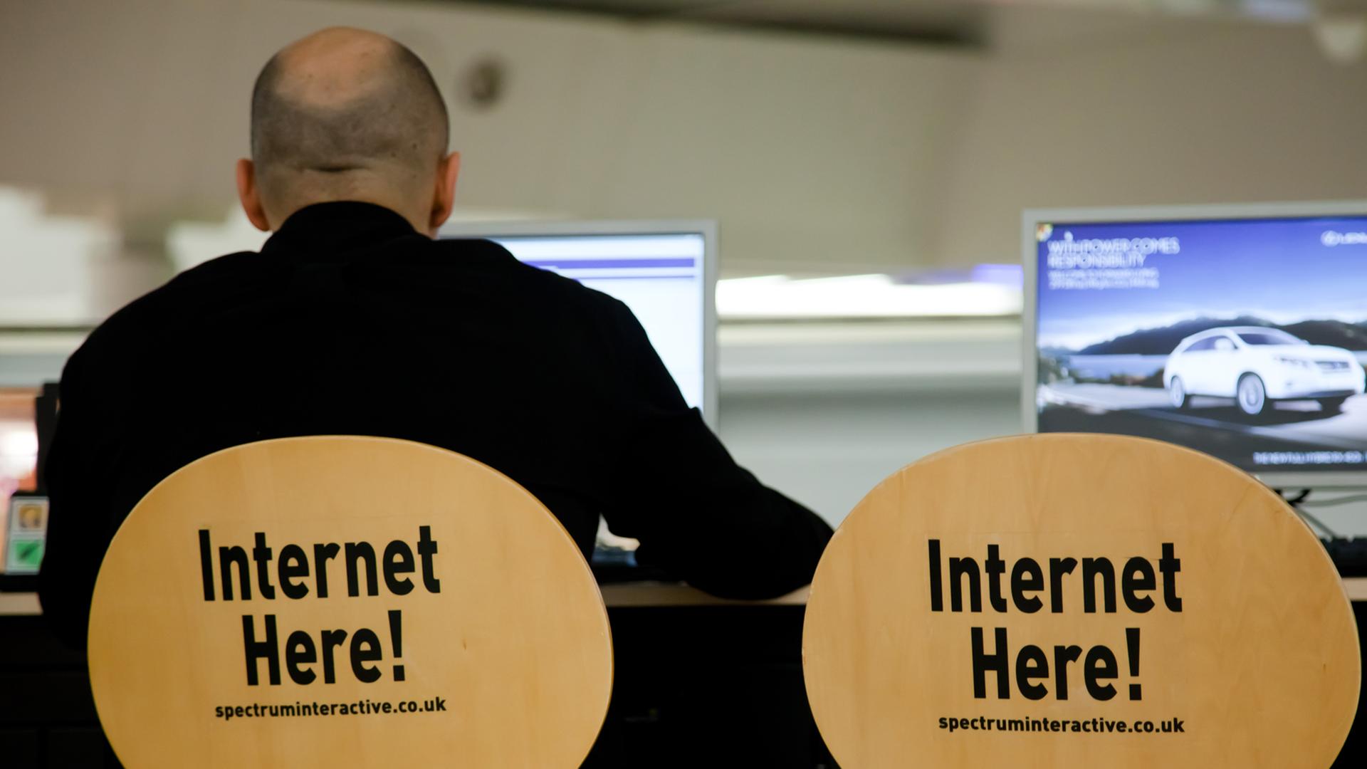 Ein Mann sitzt auf einem Stuhl, auf dessen Rückenlehne "Internet here!" steht, neben ihm ein identischer Stuhl und ein Computerbildschirm Ein Mann sitzt auf einem Stuhl, auf dessen Rückenlehne "Internet here!" steht, neben ihm ein identischer Stuhl und ein Computerbildschirm