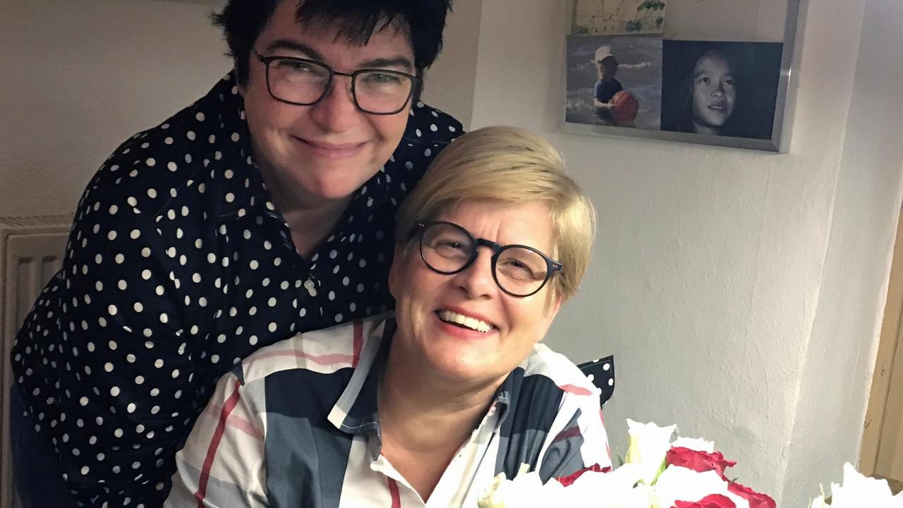 Vera (links) und Judith (rechts) Steinbeck, ein lesbisches Paar mit zwei Kindern, schaut in die Kamera. Judith hält einen Blumenstrauß in der Hand.