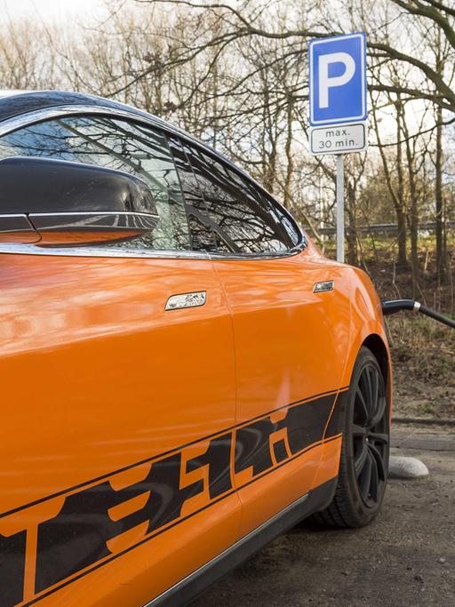 Ein orangefarbenes Elektro-Auto mit der Aufschrift "Tesla" ist an eine Ladestation angeschlossen.