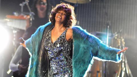 Whitney Houston auf der Bühne, lacht und streckt die Hände aus. Sie trägt ein silbernes Glitzerkleid und darüber einen grünen Pelzmantel.