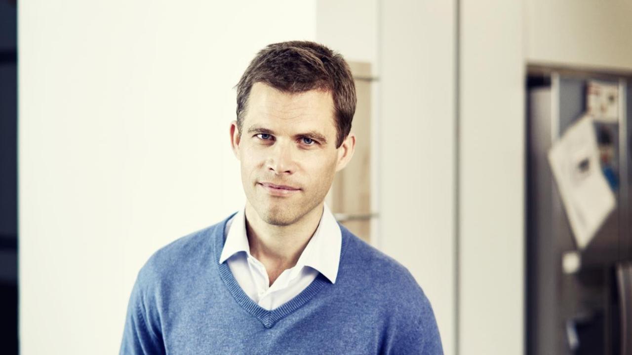 Bas Kast steht vor einer weißen Wand und trägt über einem weißen Hemd einen blauen Pullover. Bas Kast steht vor einer weißen Wand und trägt über einem weißen Hemd einen blauen Pullover.