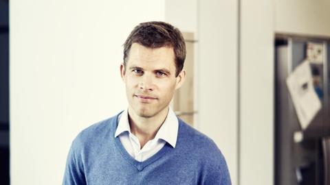 Bas Kast steht vor einer weißen Wand und trägt über einem weißen Hemd einen blauen Pullover.
