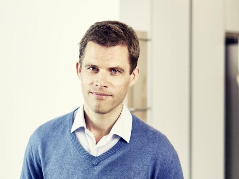 Bas Kast steht vor einer weißen Wand und trägt über einem weißen Hemd einen blauen Pullover.