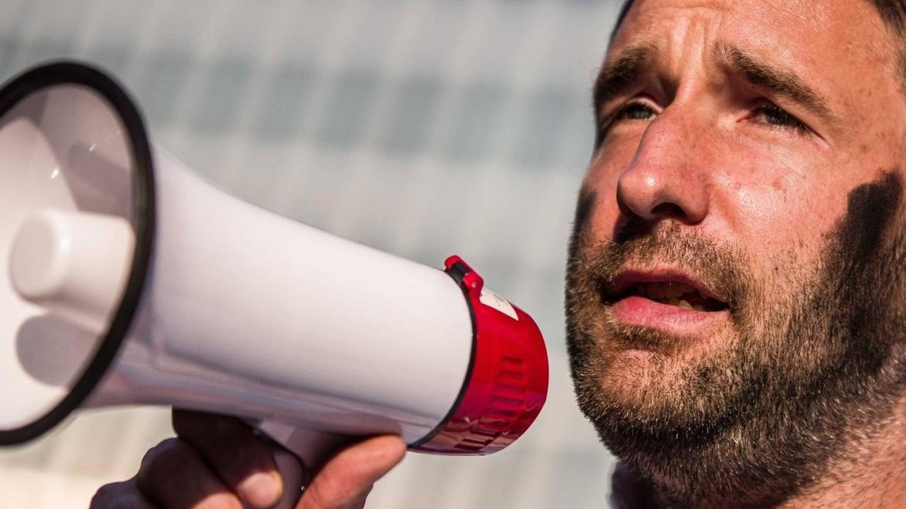Ruch spricht in ein Megaphon. Sein Gesicht ist teilweise schwarz angemalt. Ruch spricht in ein Megaphon. Sein Gesicht ist teilweise schwarz angemalt.