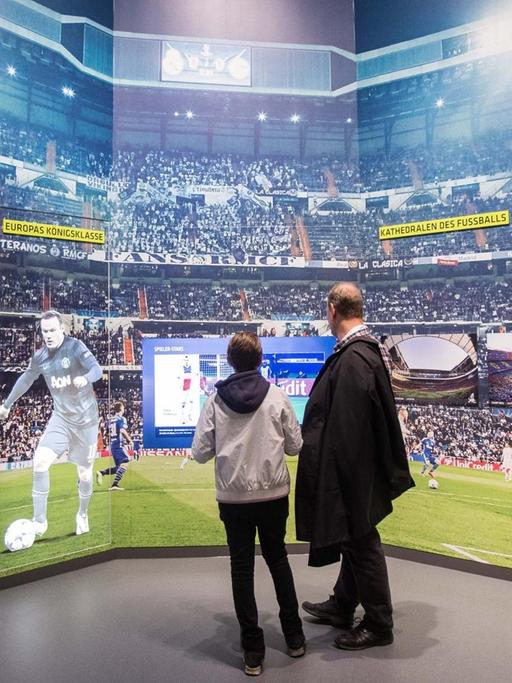 Ein Mann und ein Kind schauen auf einem Monitor ein Fußpballspiel an. Der Monitor befindet sich in einer Wand mit der Darstellung eines Stadion-Innenraums. Auf der linken Seite sind Ganzkörperportraits der Spieler Ronaldo, Robben und Rooney zu sehen.