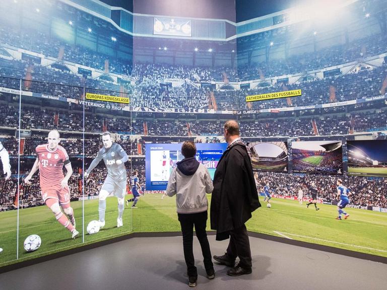 Ein Mann und ein Kind schauen auf einem Monitor ein Fußpballspiel an. Der Monitor befindet sich in einer Wand mit der Darstellung eines Stadion-Innenraums. Auf der linken Seite sind Ganzkörperportraits der Spieler Ronaldo, Robben und Rooney zu sehen.