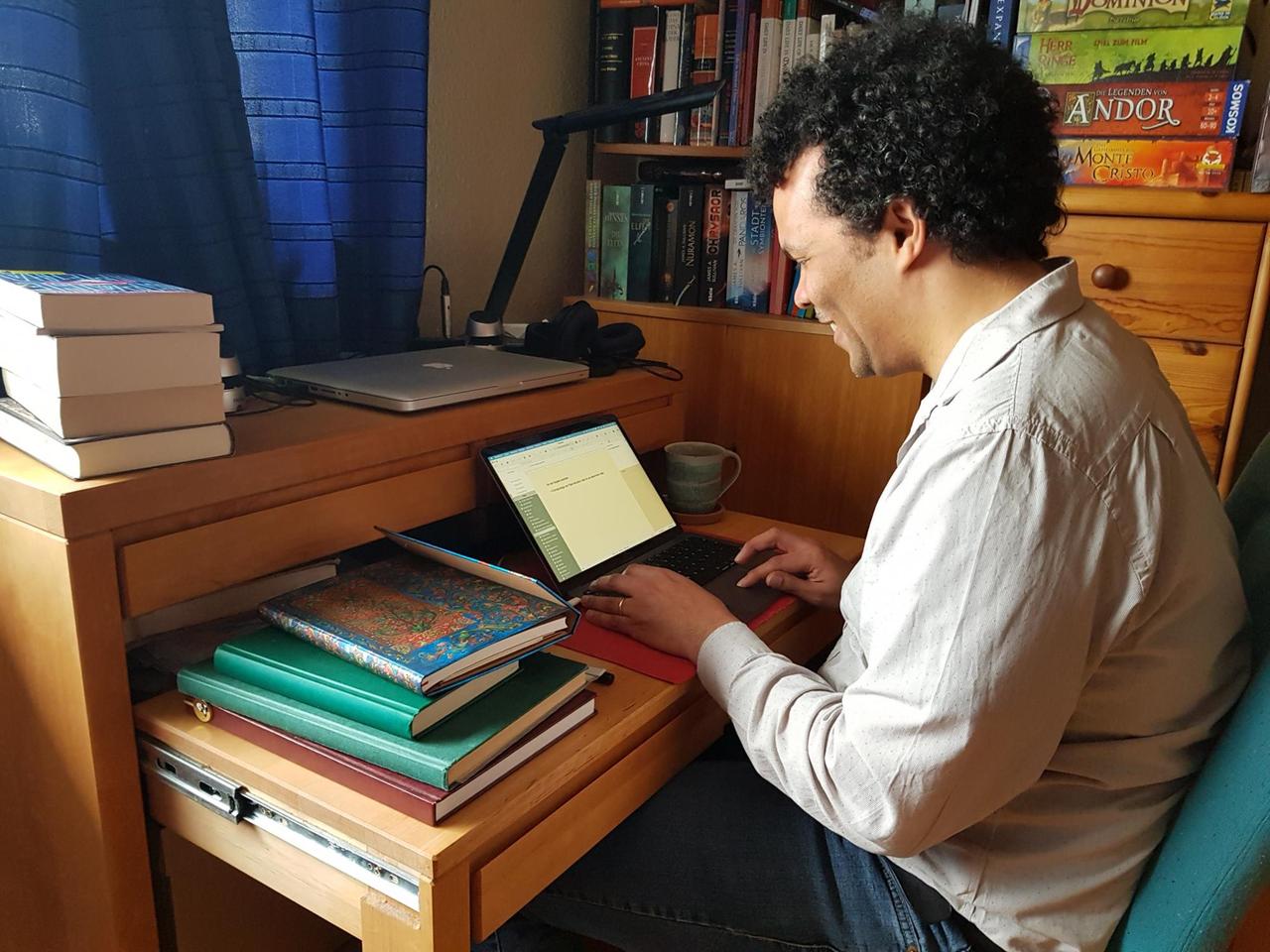 James A. Sullivan sitzt an seine Schreibtisch und blickt in seinen Laptop. Er lächelt dabei. Um ihr herum liegen viele Bücher gestapelt. In dem Regal hinter ihm stehen weitere Bücher und Spiele. James A. Sullivan sitzt an seine Schreibtisch und blickt in seinen Laptop. Er lächelt dabei. Um ihr herum liegen viele Bücher gestapelt. In dem Regal hinter ihm stehen weitere Bücher und Spiele.