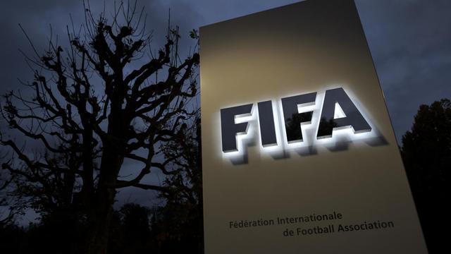 Eine Stele mit dem leuchtenden FIFA-Schriftzug in der späten Abenddämmerung, dahinter ein kahler Baum.