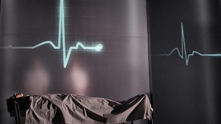 Ein Mensch liegt auf einem Krankenhausbett und blickt nach oben. Im Hintergrund sein visualisierter Herzschlag.