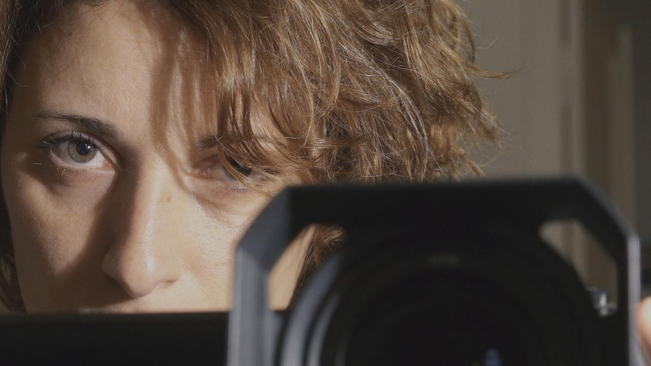 Ein Frau mit braunen, lockigen Haaren schaut über ein Kamerobjektiv.