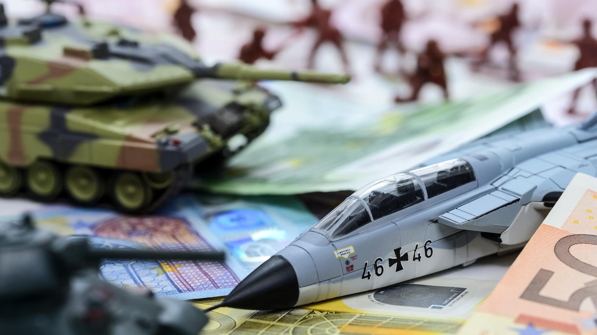 Das Symbolbild zeigt das Modell eines Panzers und eines Militärjets sowie Geldscheine.
