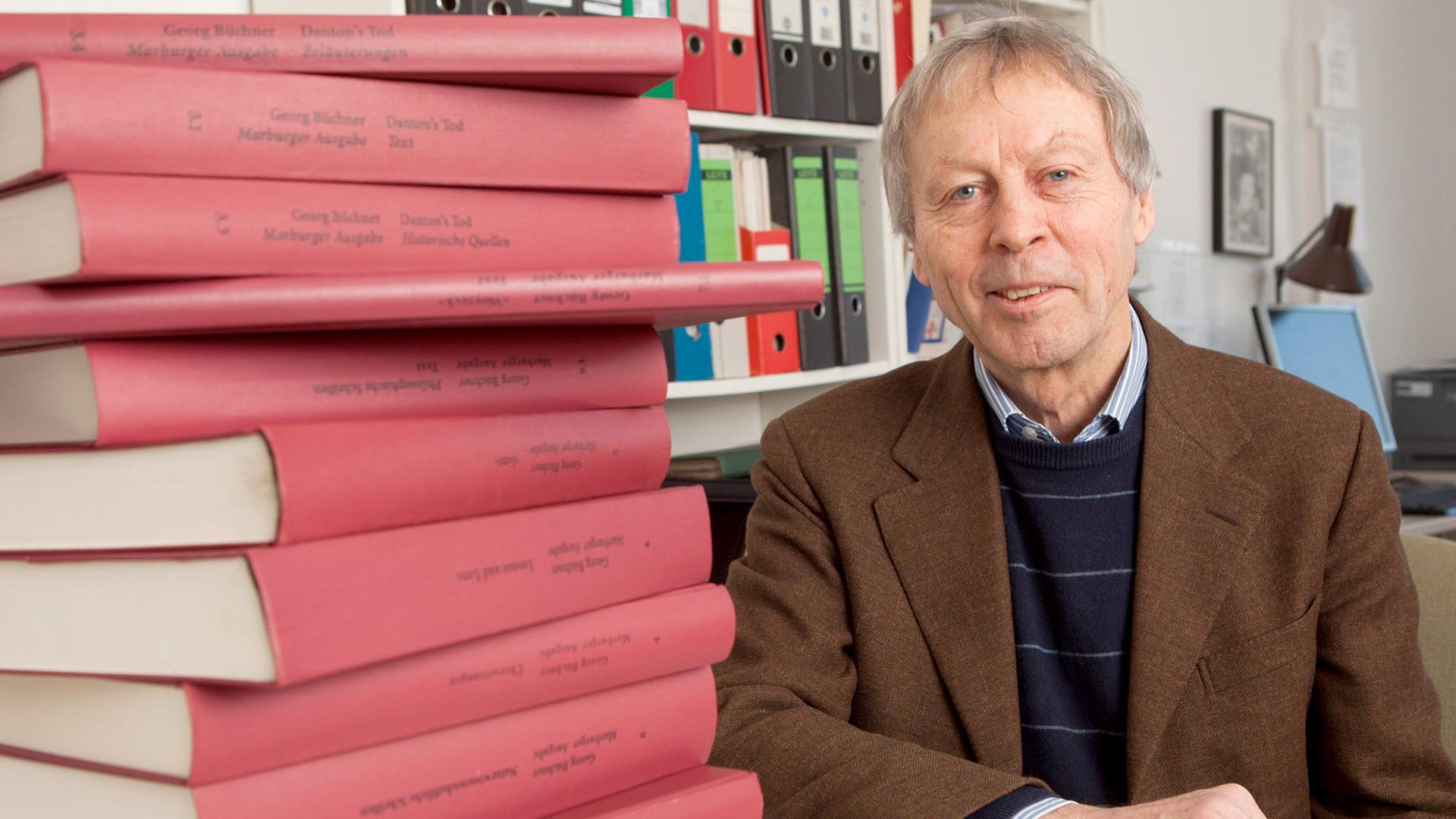 Ein Mann sitzt an einem runden Tisch, vor ihm ein großer Stapel rot eingebundener Bücher, hinter ihm weitere in einem Regal. Ein Mann sitzt an einem runden Tisch, vor ihm ein großer Stapel rot eingebundener Bücher, hinter ihm weitere in einem Regal.