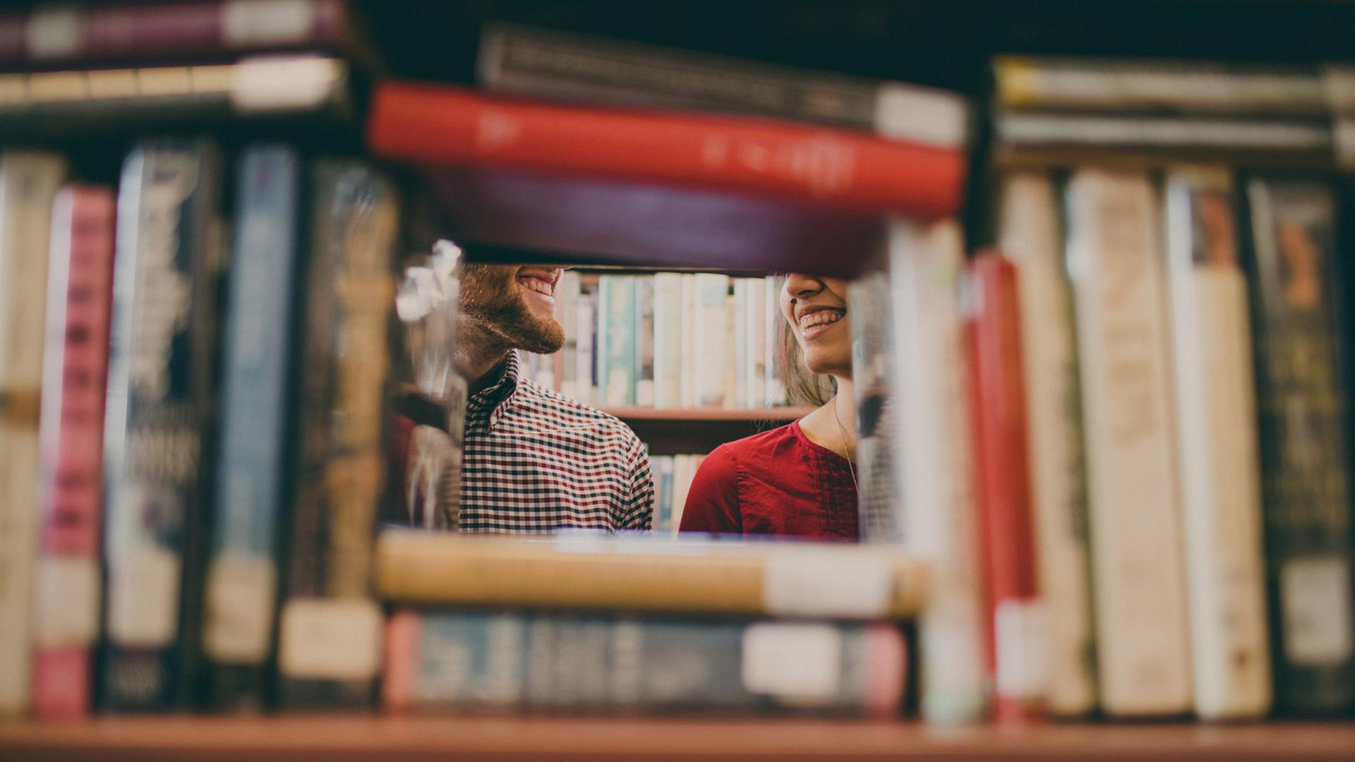 Ein Mann und eine Frau stehen zwischen Bücherregalen und lächeln sich an. Ein Mann und eine Frau stehen zwischen Bücherregalen und lächeln sich an.