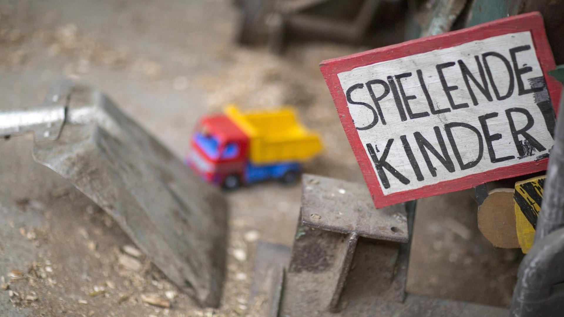 Ein Schild mit der Aufschrift "Spielende Kinder" an einem Spielplatz, im Hintergrund ein Spielzeug-LKW Ein Schild mit der Aufschrift "Spielende Kinder" an einem Spielplatz, im Hintergrund ein Spielzeug-LKW