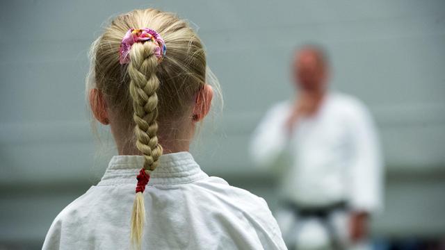 Ein kleines Mädchen mit einem blonden Zopf, von hinten aufgenommen, lauscht den Erklärungen ihres Karate Trainers.