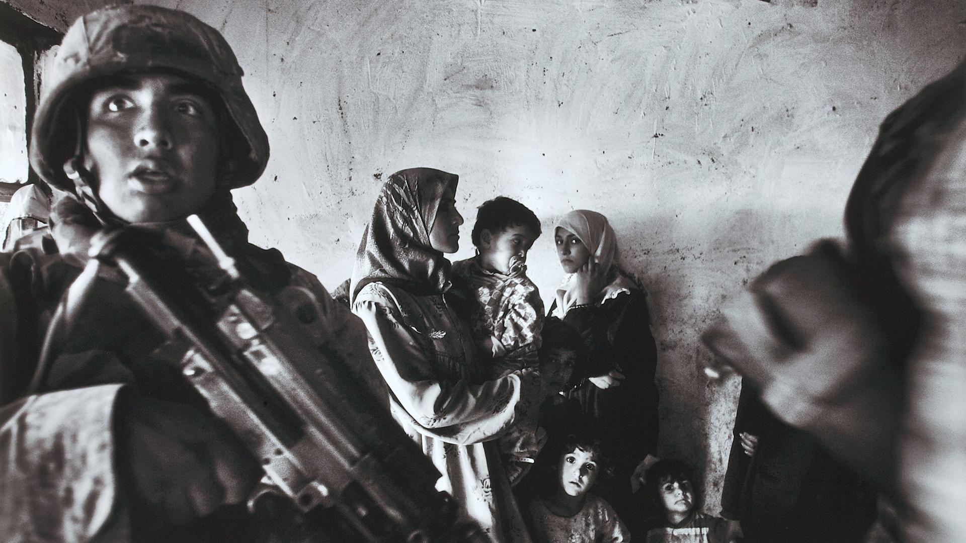 Ein Schwarz-weiß-Foto von Anja Niedringhaus: US-Marineinfanteristen mit Gewehren führen Ende 2004 eine Razzia im Haus eines irakischen Abgeordneten im Stadtteil Abu Ghraib durch. Zwei Frauen mit Kopftuch stehen mit mehreren Kindern an der Wand.