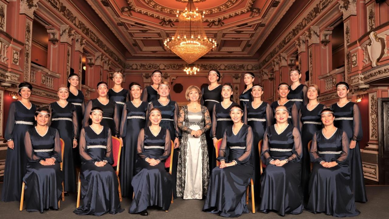 Der Chor sitzt in dunkelblauen Kleidern und zarten Stirnbändern einem historischen Raum mit schwerer, dekorierter Holzdecke. Der Chor sitzt in dunkelblauen Kleidern und zarten Stirnbändern einem historischen Raum mit schwerer, dekorierter Holzdecke.