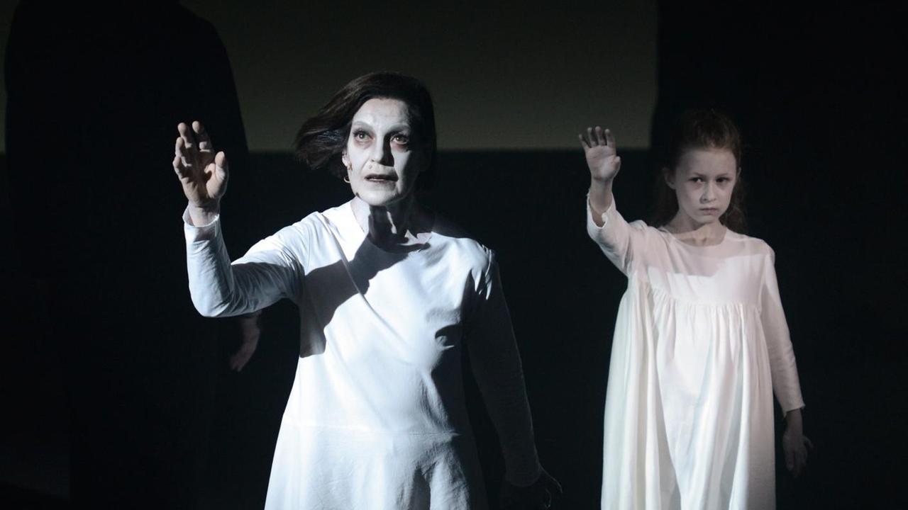 Eine Frau und ein Mädchen in weißen langen Kleidern stehen angestrahlt auf sonst dunkler Bühne und heben die Arme.