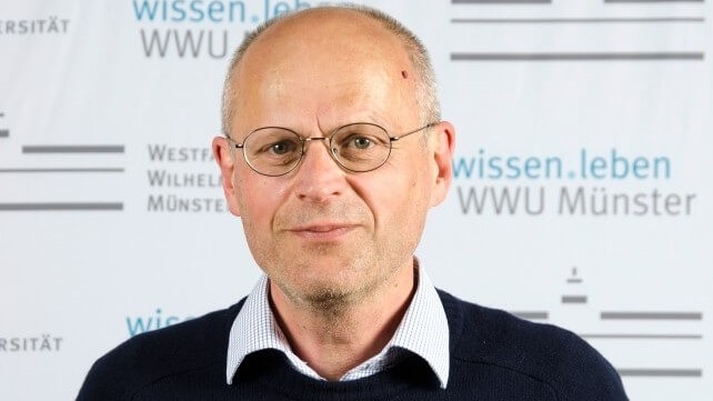 Ein Mann mit Brille, in Hemd und Pullover, steht vor einem Plakat der Universität Münster und schaut leicht stirnrunzelnd, aber lächelnd in die Kamera. Ein Mann mit Brille, in Hemd und Pullover, steht vor einem Plakat der Universität Münster und schaut leicht stirnrunzelnd, aber lächelnd in die Kamera.