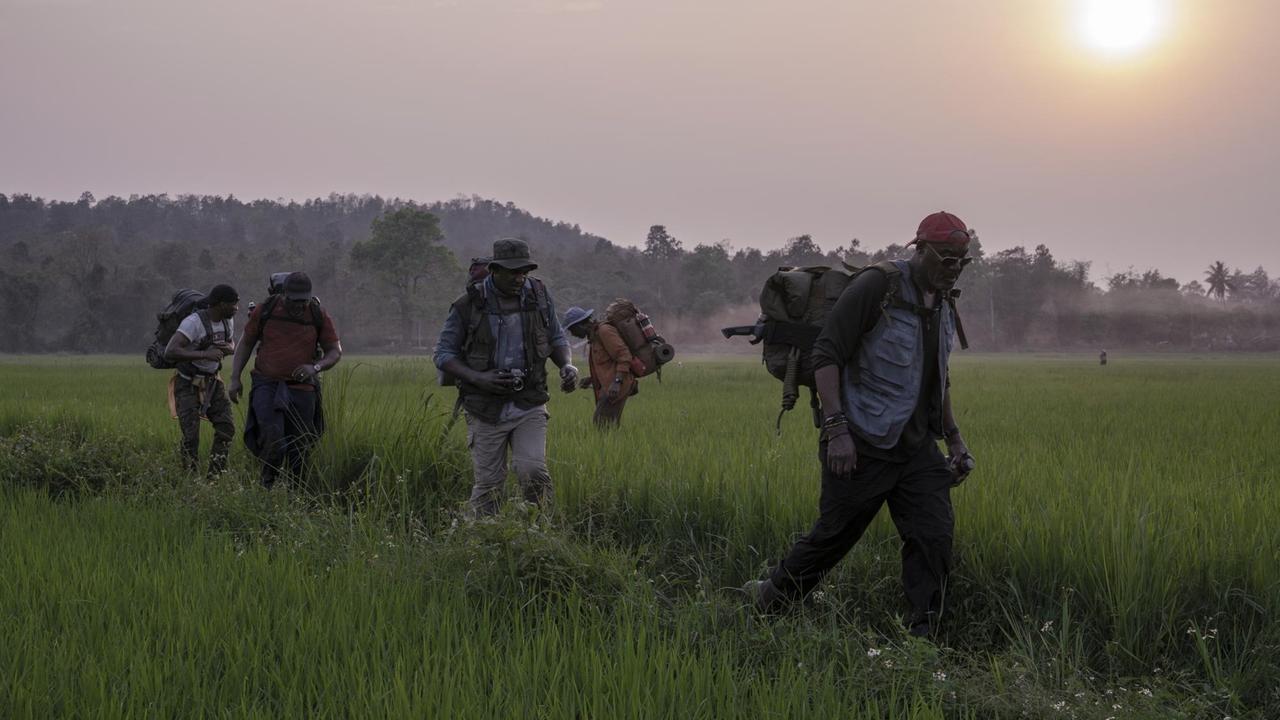 5 Männer mit großen Rucksäcken laufen in der Abenddämmerung durch ein Reisfeld.