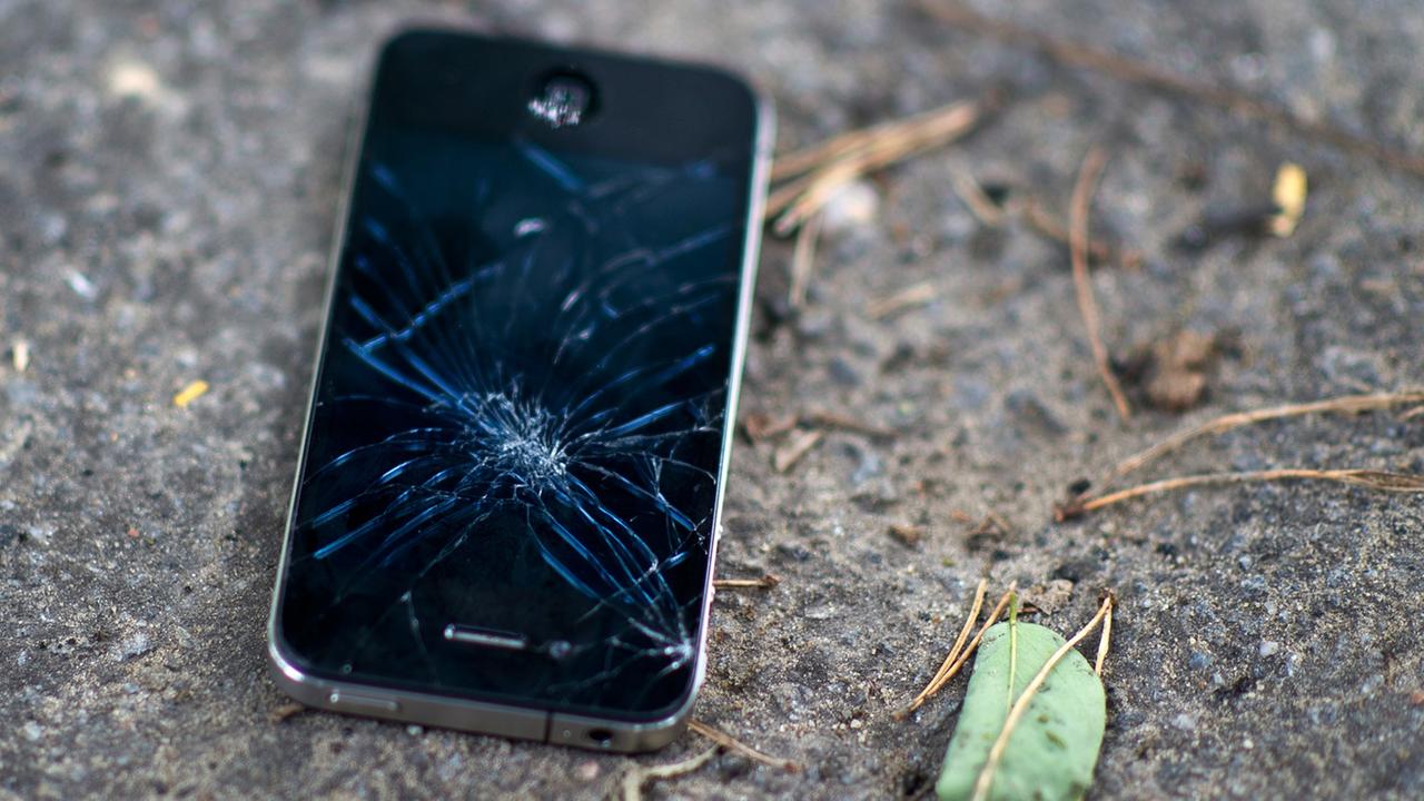 Ein schwarzes Mobiltelefon liegt mit zersplittertem Display auf dem Boden. Ein schwarzes Mobiltelefon liegt mit zersplittertem Display auf dem Boden.