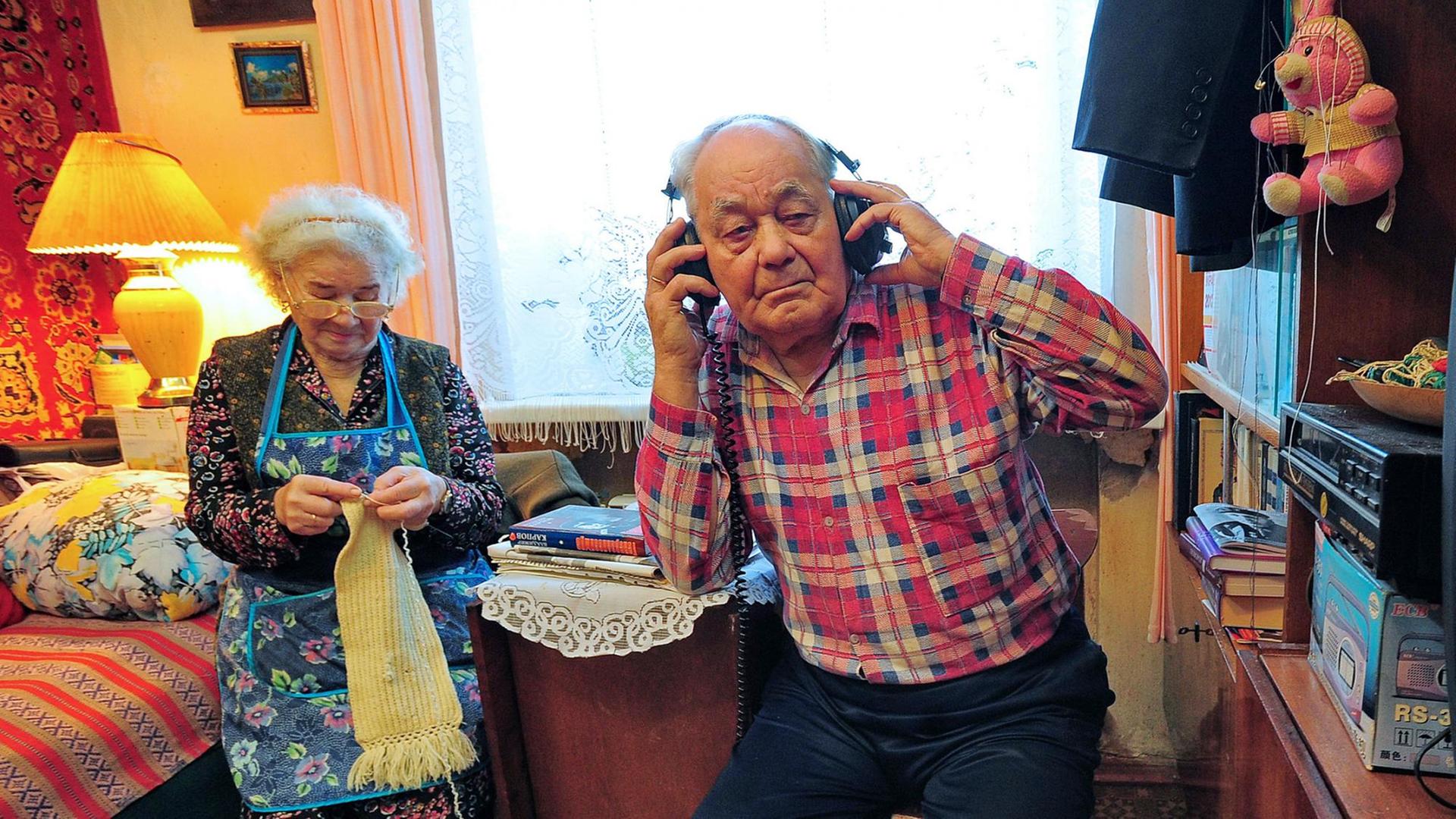 Ein älteres Ehepar in seinem Wohnzimmer: Sie strickt, er hat einen Kopfhörer auf.