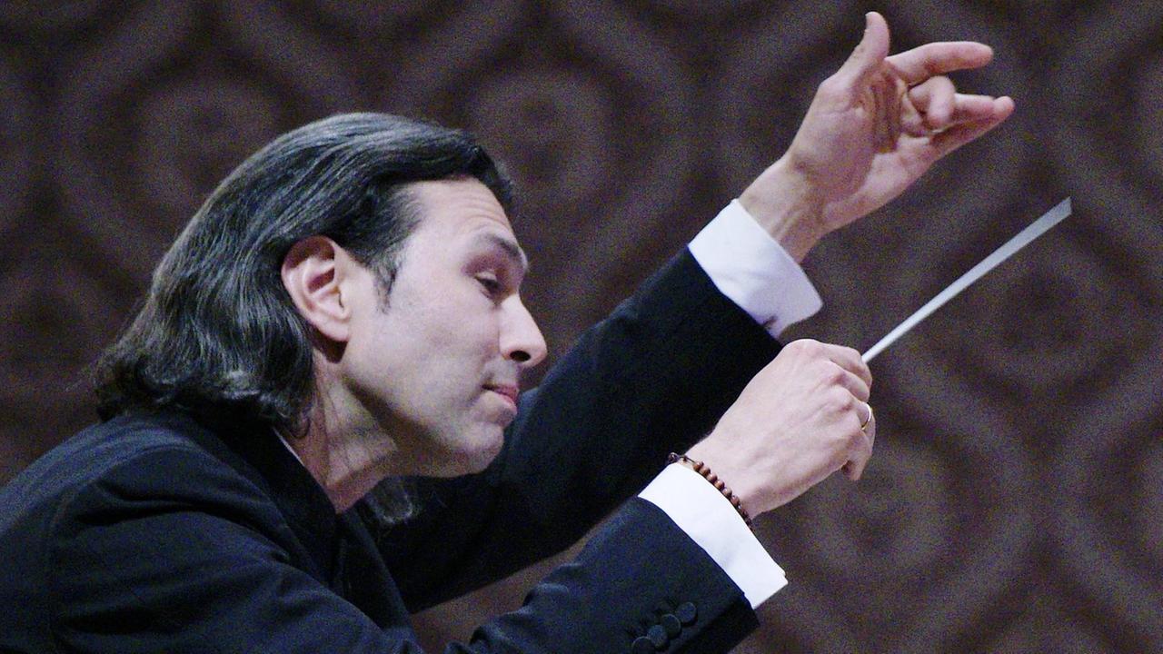 Der Dirigent im Profil, beide Hände für das Orchester erhoben. Der Dirigent im Profil, beide Hände für das Orchester erhoben.