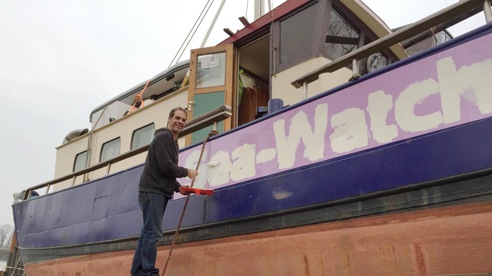 Harald Höppner steht auf einem Holzbrett vor einem Schiff, mit Malerutensilien in der Hand, auf dem Schiff steht "Sea-Watch" geschrieben.