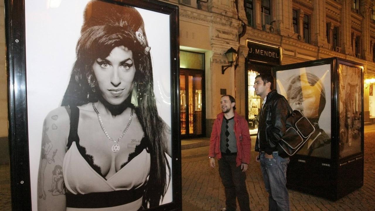 Zwei Passanten stehen vor einem Schaukasten, in dem ein Porträt von Amy Winehouse gezeigt wird. Zwei Passanten stehen vor einem Schaukasten, in dem ein Porträt von Amy Winehouse gezeigt wird.