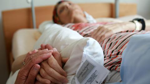 Eine ehrenamtliche Helferin hält die Hand einer sterbenden Bewohnerin, die in ihrem Bett im Christophorus Hospiz in München liegt.