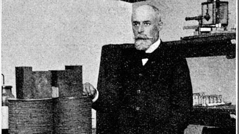 Ein zeitgenössisches Schwarz-Weiß-Foto zeigt den Physiker Antoine Henri Becquerel In Anzug ins seinem Labor an einem Tisch stehend auf dem sich zwei große Spulen befinden. Ein zeitgenössisches Schwarz-Weiß-Foto zeigt den Physiker Antoine Henri Becquerel In Anzug ins seinem Labor an einem Tisch stehend auf dem sich zwei große Spulen befinden.